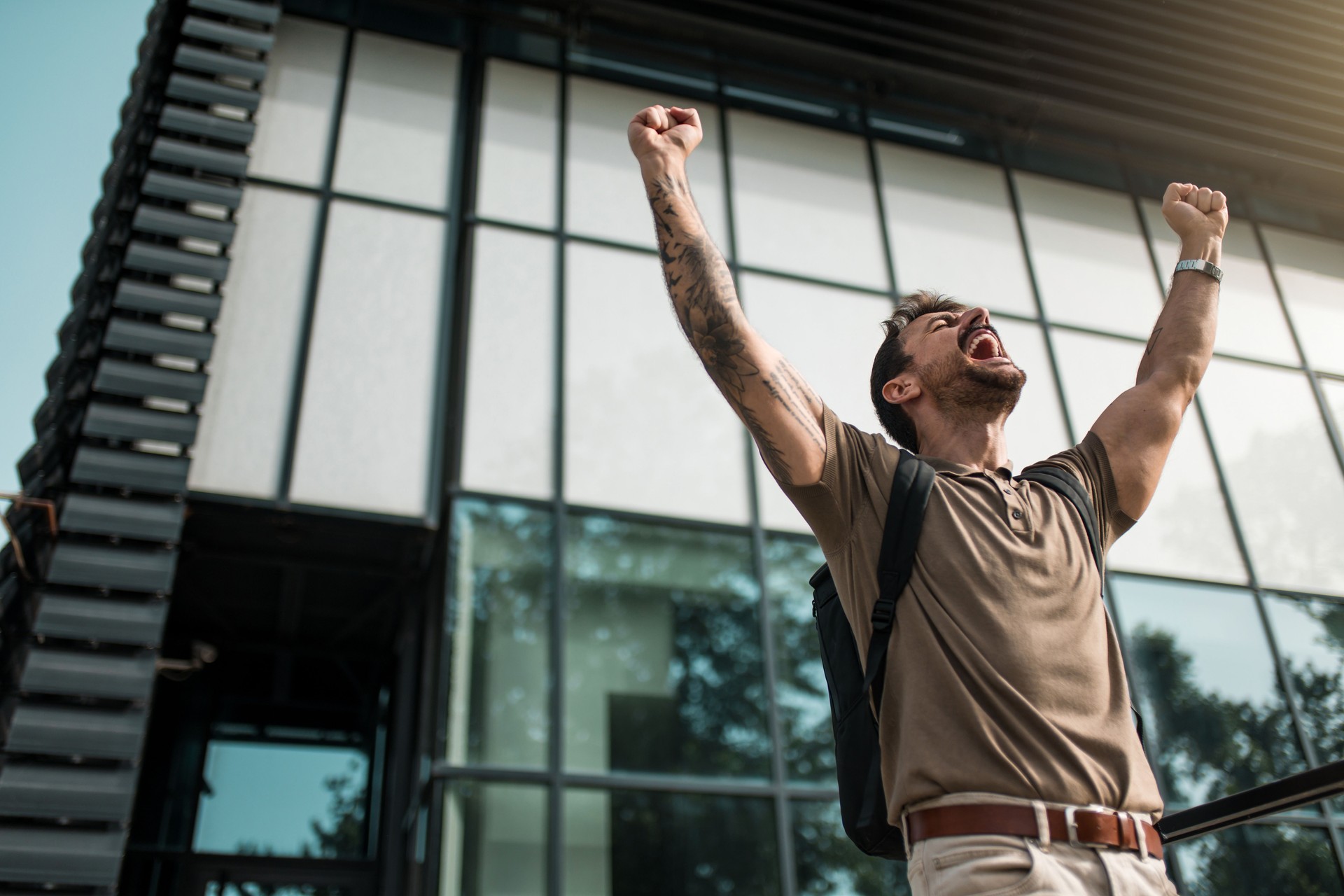 Joven extasiado celebrando el éxito mientras está de pie frente a un edificio de oficinas.